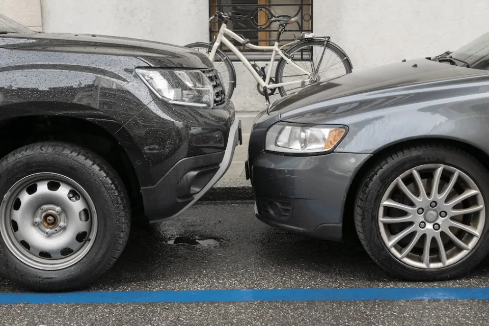 Two grey cars parked nose to nose, one taller than the other, on city curb with parked bicycle visible on sidewalk.